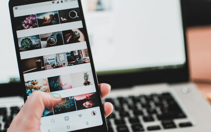 A person uses a smartphone to browse social media in front of a laptop, emphasizing modern technology and lifestyle.