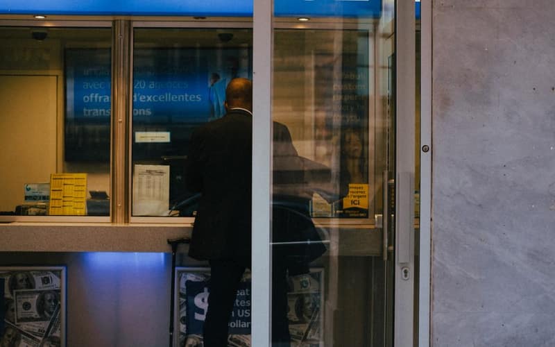 Man at a currency exchange office window, showing currency rates inside a bustling city.