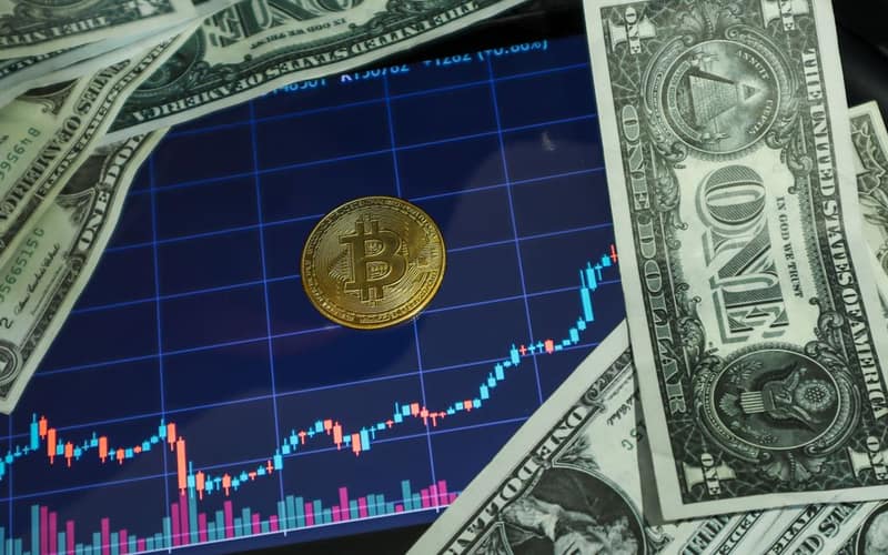 Bitcoin coin on a tablet showing stock chart, surrounded by dollar bills.