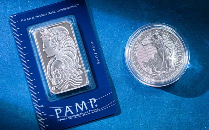 Close-up of a silver coin and a PAMP bar on a blue textured background emphasizing wealth.