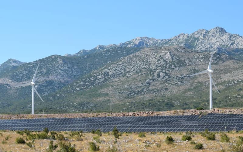 A scenic view of windmills and solar panels against mountainous terrain under a clear blue sky.