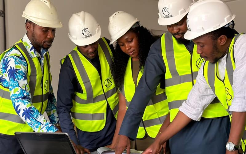 A diverse team of five engineers in safety gear examines construction plans indoors.