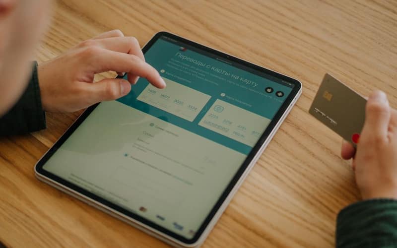 Person making an online payment using a tablet and credit card on a wooden desk.