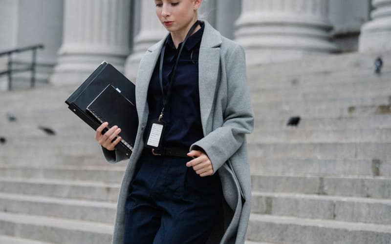 Young stylish female lawyer in formal clothes with name tag holding folders while going downstairs near stone building with columns