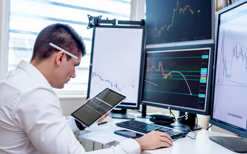 Professional analyzing stock market graphs on multiple monitors at work desk.