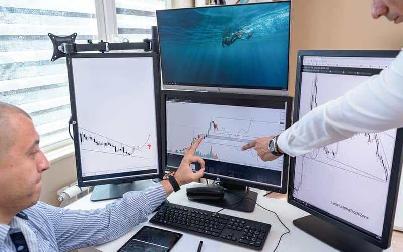Two business professionals analyzing financial data on multiple computer screens in an office setting.
