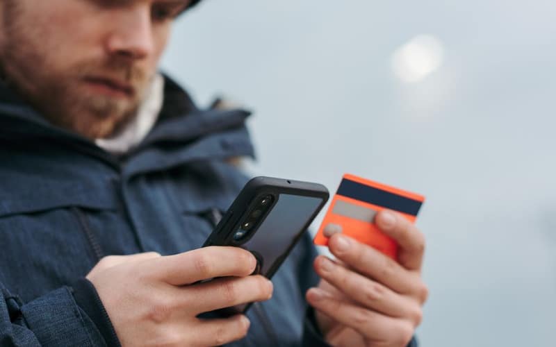 Crop male in outwear entering details of credit card on mobile phone while making online payment for purchase in daytime
