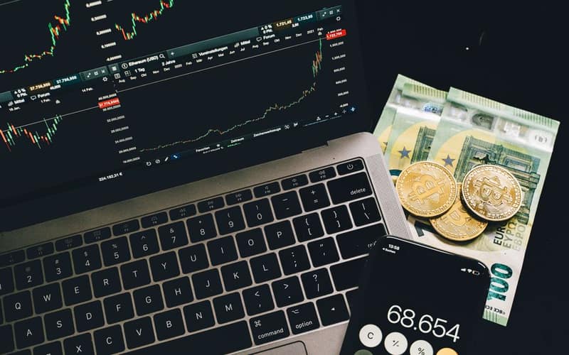 A laptop displaying stock charts with Bitcoin, Euros, and a cellphone calculator, showcasing financial analysis.