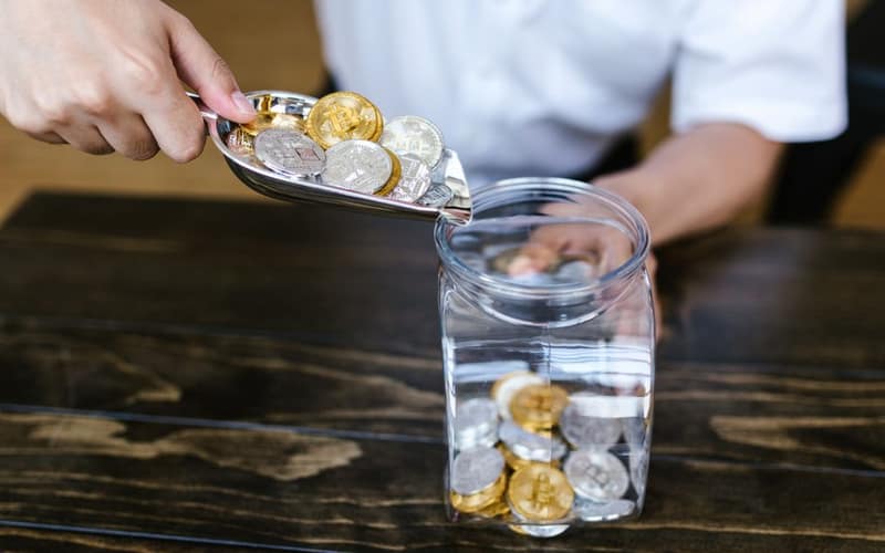 Hand placing gold and silver coins into a glass jar on a wooden table indoors.